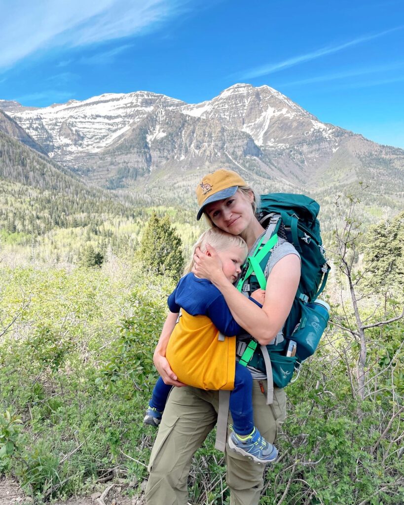 Mother holding son in child carrier on hiking trail. Son is wearing merino wool outfit